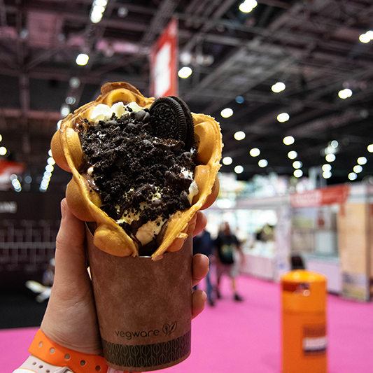 A photo of an oreo waffle dessert being held up with convention stalls in the background.