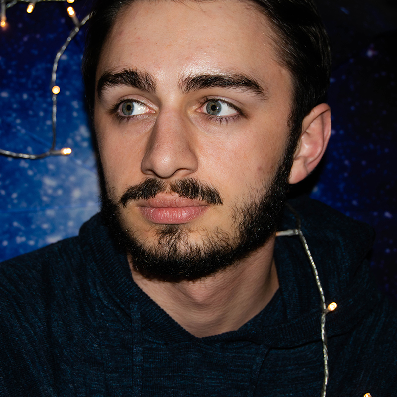 A close up photo of a man staring behind the camera in front of a starry blue backdrop with LED lights.